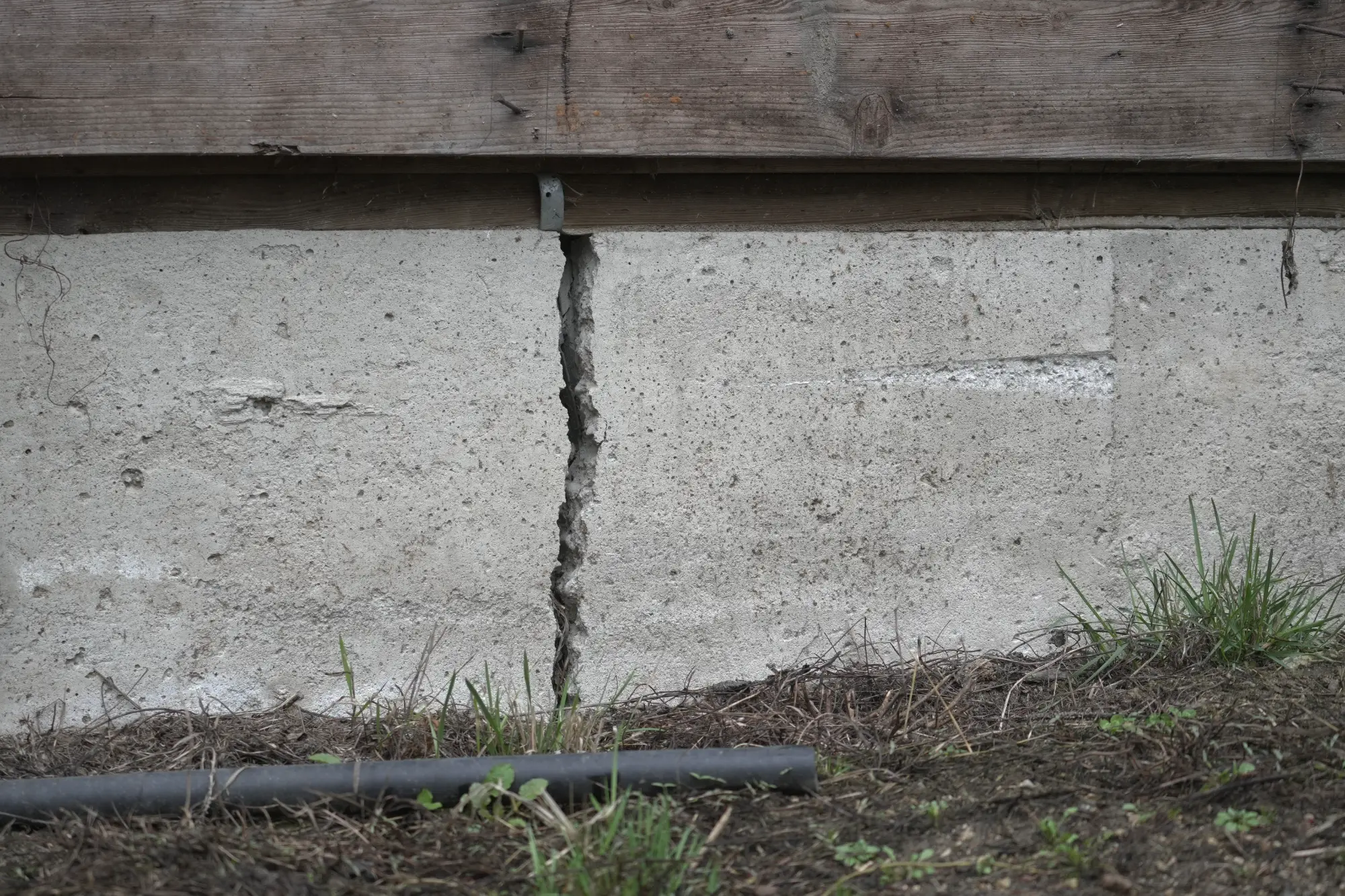 A large vertical crack runs through a concrete foundation wall beneath a wooden structure, with soil, grass, and a black pipe visible at the base.