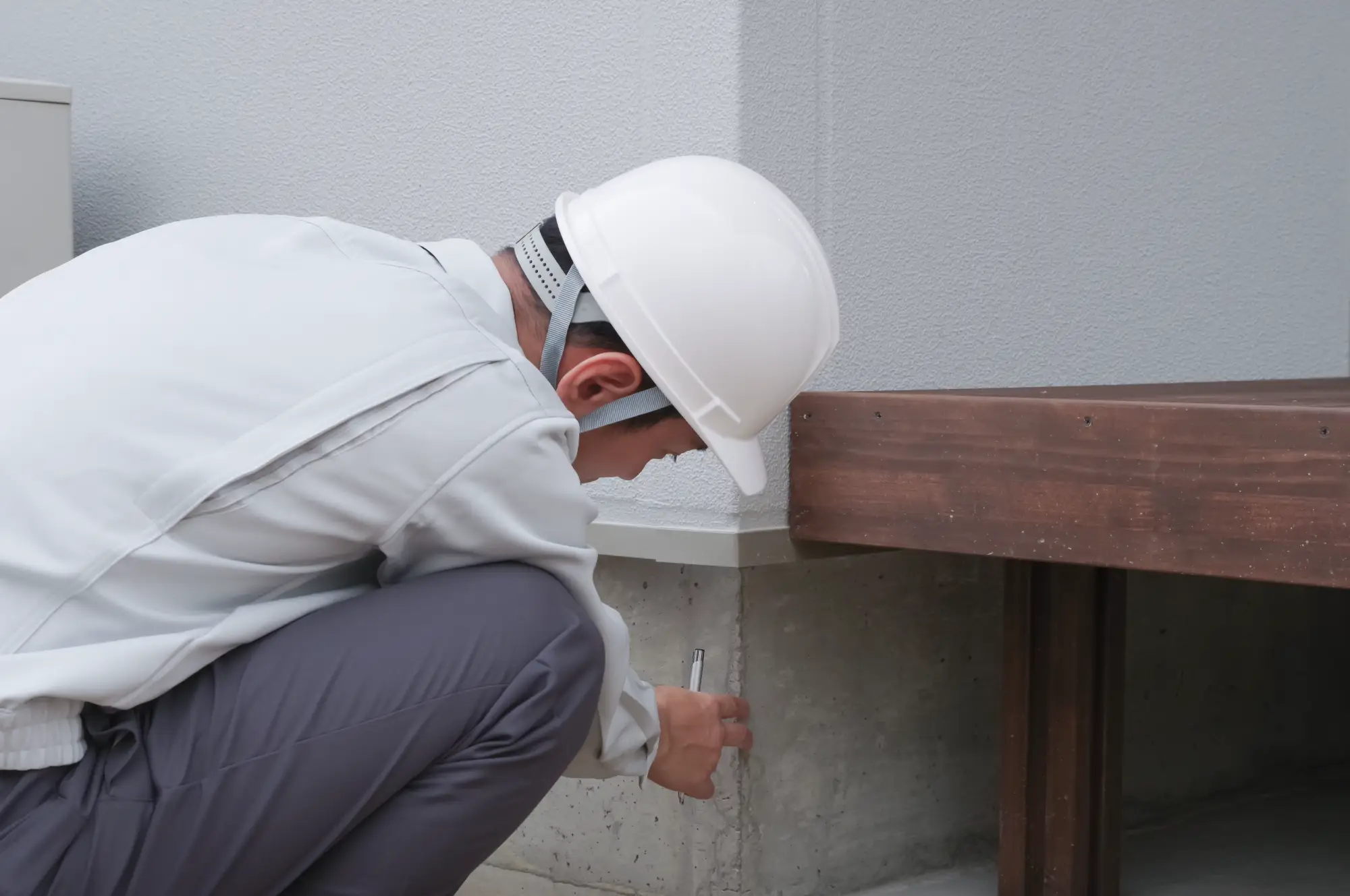 A person wearing a white hard hat and work clothes inspects the foundation of a building, closely examining or measuring a crack in the concrete wall near a wooden structure.