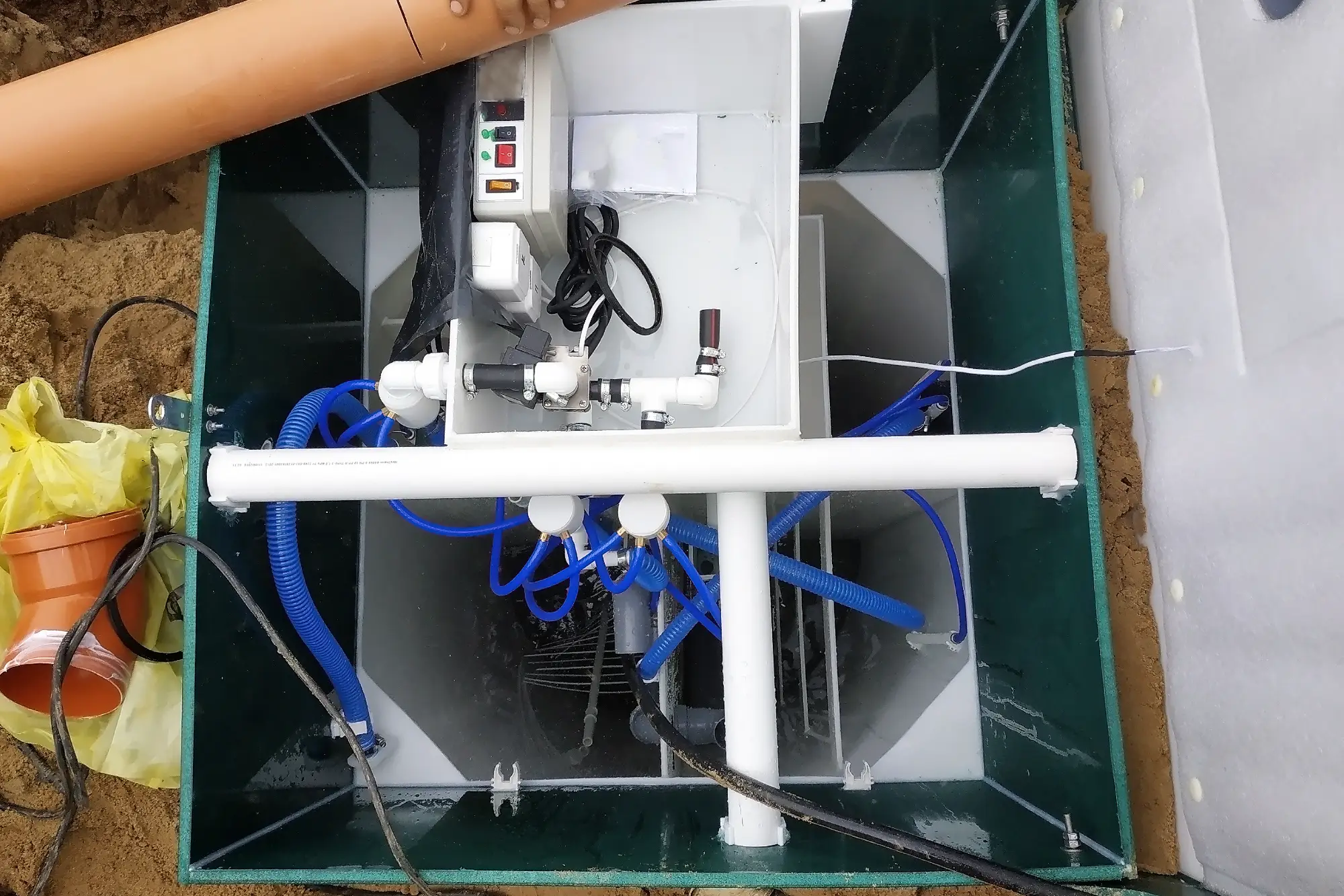 A close up view of a wastewater treatment system installed in the ground, showing pipes, electrical components, blue hoses, and control panel inside a rectangular tank.