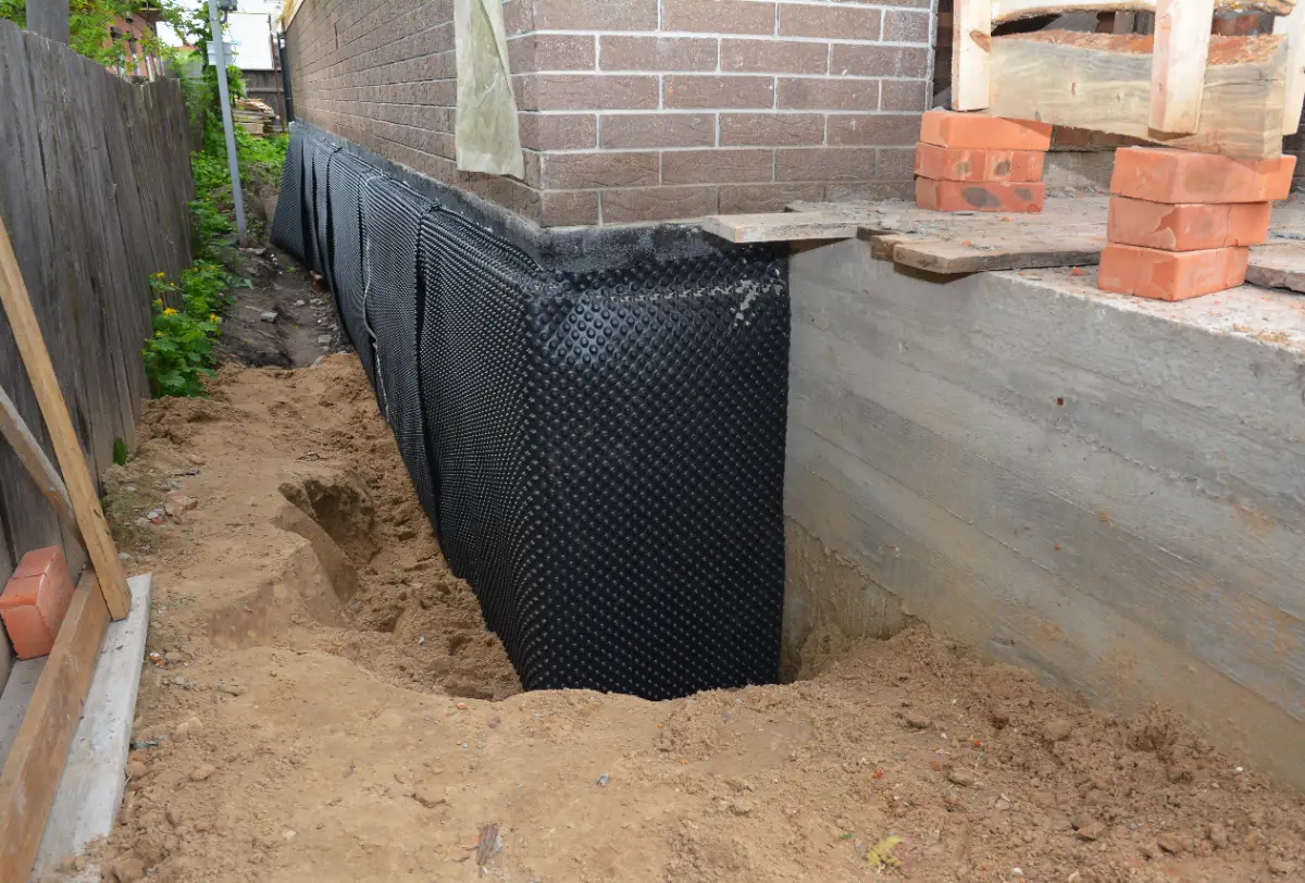 A house foundation is partially exposed with black waterproofing membrane applied to the concrete wall. Sand and dirt are piled beside the foundation, and construction materials are visible nearby.