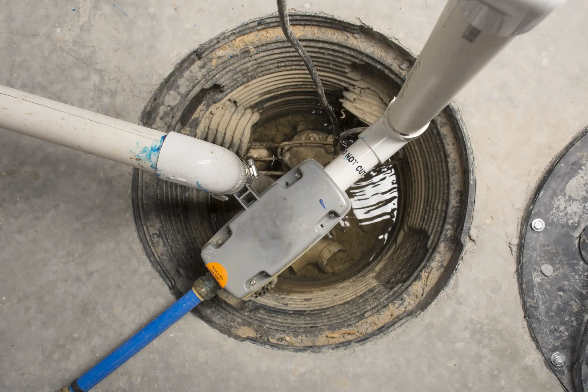 A close up view of a sump pump installed in a circular pit in a concrete floor, with several white and blue pipes connected and some standing water visible in the pit.