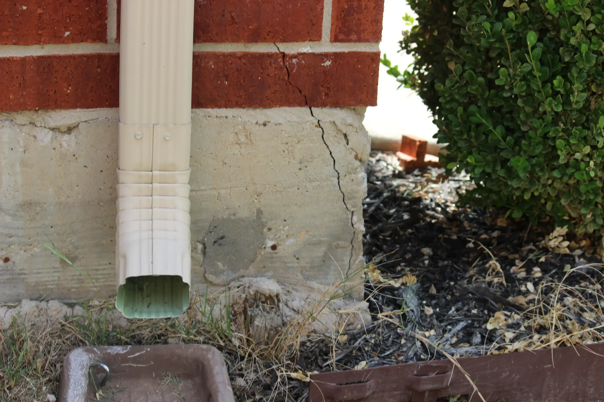 A cracked concrete foundation beneath a brick wall near a downspout, with dry grass, mulch, and a shrub nearby.